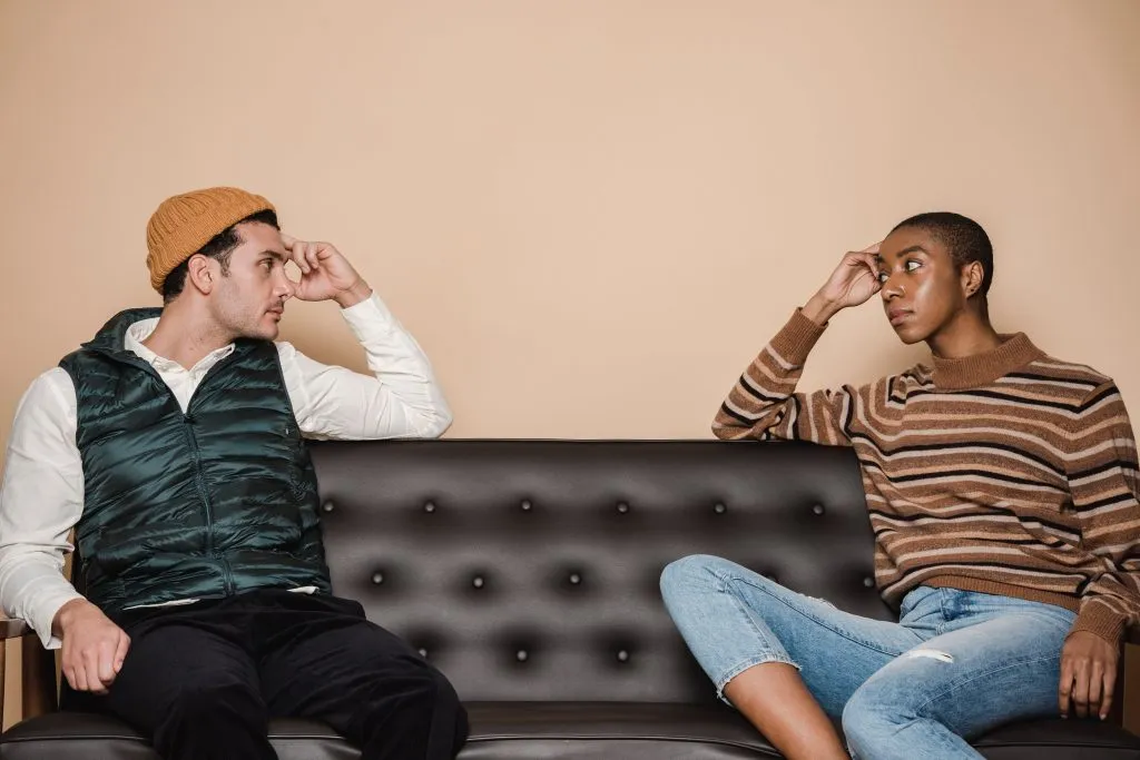 A white man and a black woman sit on opposite sides of a couch and look at each other unhappily, illustrating that Susan Gonzales helps couples understand their attachment styles.