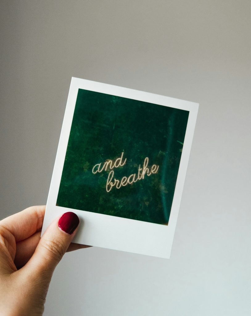 hand holding a polaroid picture of a neon sign that states "and breathe"